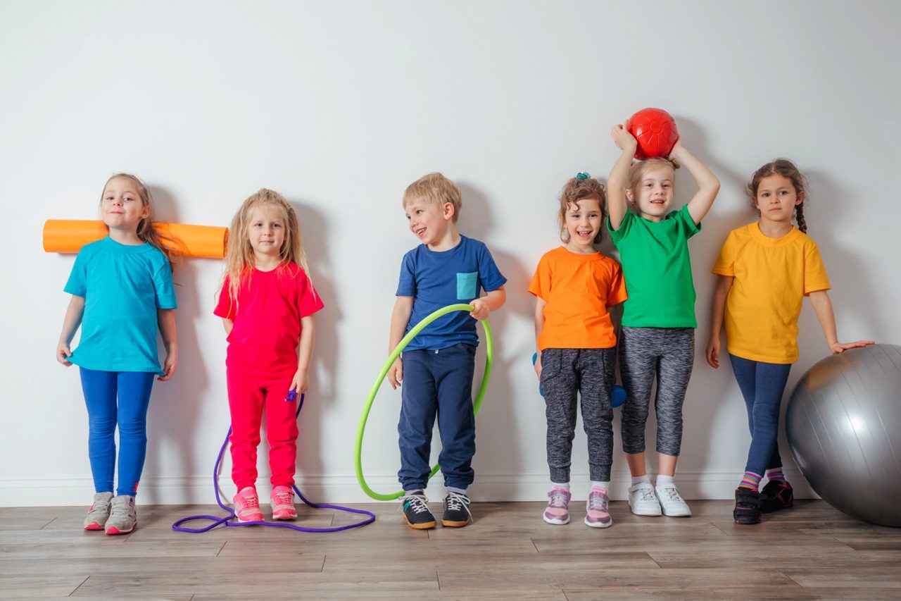 Group of cheerful young kids in colorful and comfortable sportswear posing next to white wall each holding one item of sport equipment. Active kids look tired after training.